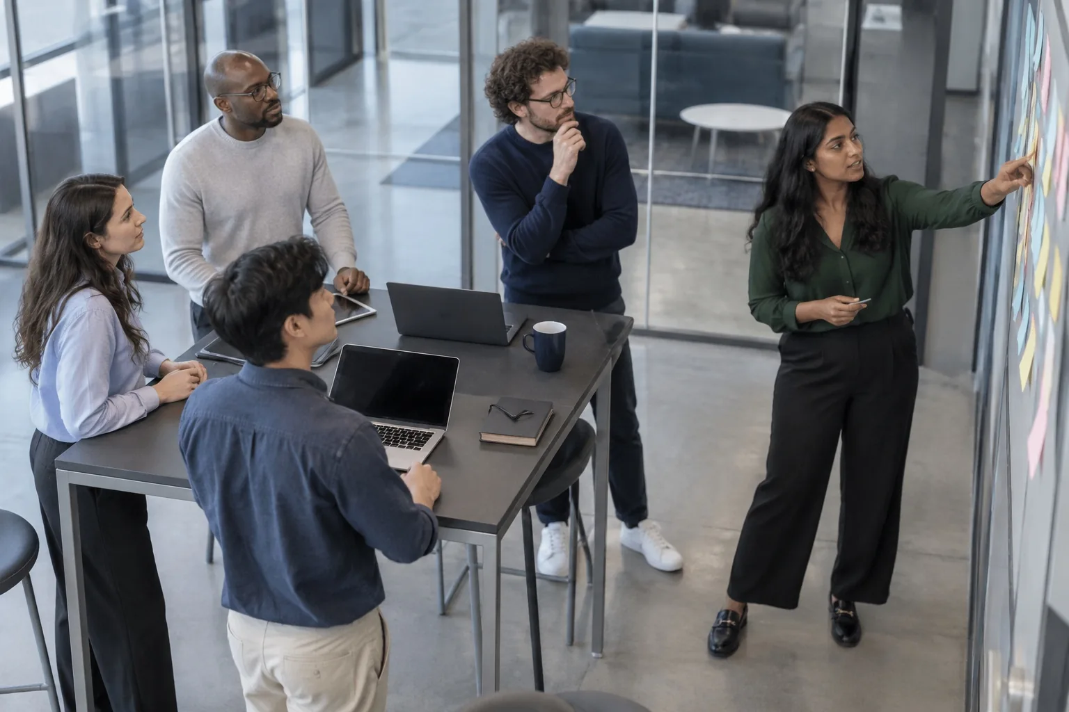 A team stands around a high table as a colleague points to a wall of sticky notes during a morning stand-up